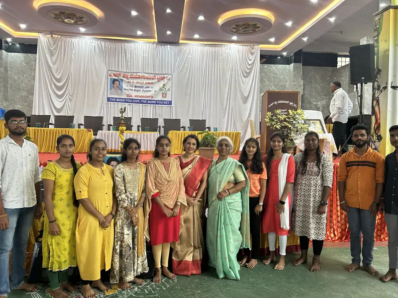 Sujatha R Shetty with students and guests in front of the Trust banner that reads The more you give, the more you get