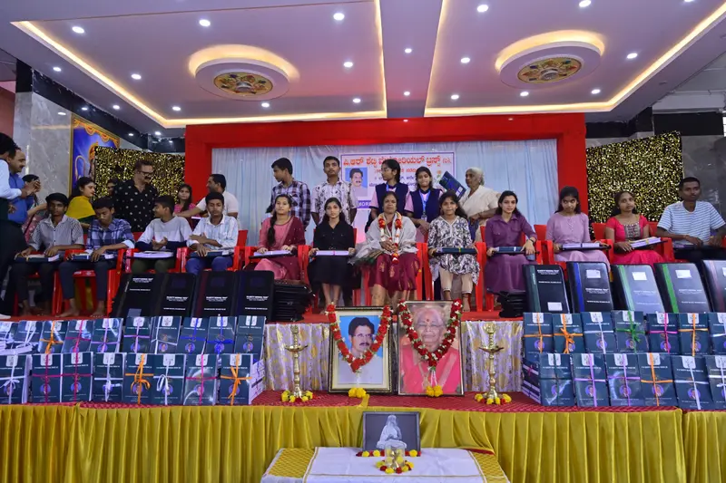 Students on stage at the Trust's Annual Donation Event, holding their new portfolios, with garlanded portraits, including P R Shetty's, in front